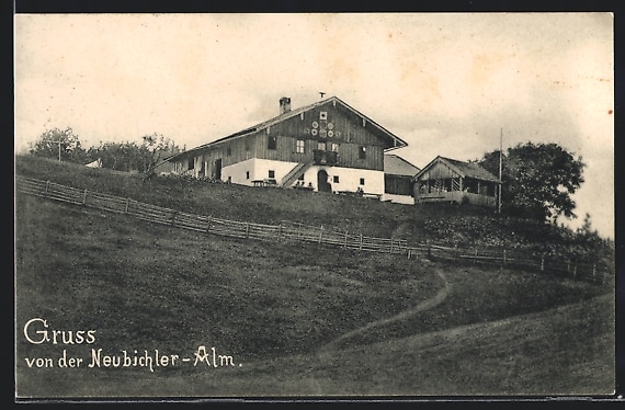 Altes Schwarzweißfoto einer Alm auf einem Hang mit Fachwerkbauernhaus, Zäunen und beschriftetem Grußtext unten links.