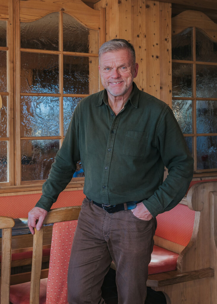 Älterer Mann mit kurzem grauem Haar und grünem Hemd steht lächelnd in holzgetäfelter Gaststube, eine Hand auf einem Stuhl.