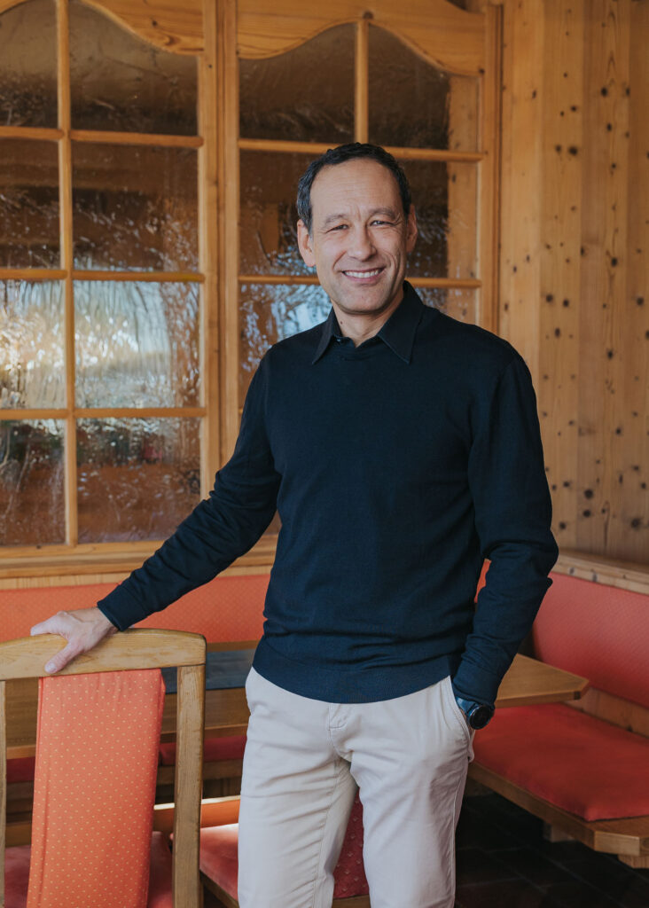 Mann mittleren Alters lächelnd in schwarzem Pullover und Hosen, steht in holzgetäfeltem Raum und lehnt an einem Stuhl.