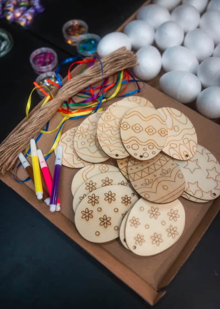 Holz-Osterei-Anhänger mit eingeritzten Blumen‑ und Musterdesigns, daneben bunte Filzstifte, Bänder, Schnur und Styroporeier.