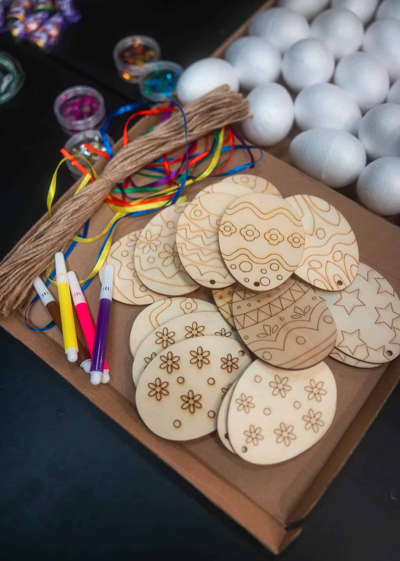 Holz-Osterei-Anhänger mit eingeritzten Blumen‑ und Musterdesigns, daneben bunte Filzstifte, Bänder, Schnur und Styroporeier.