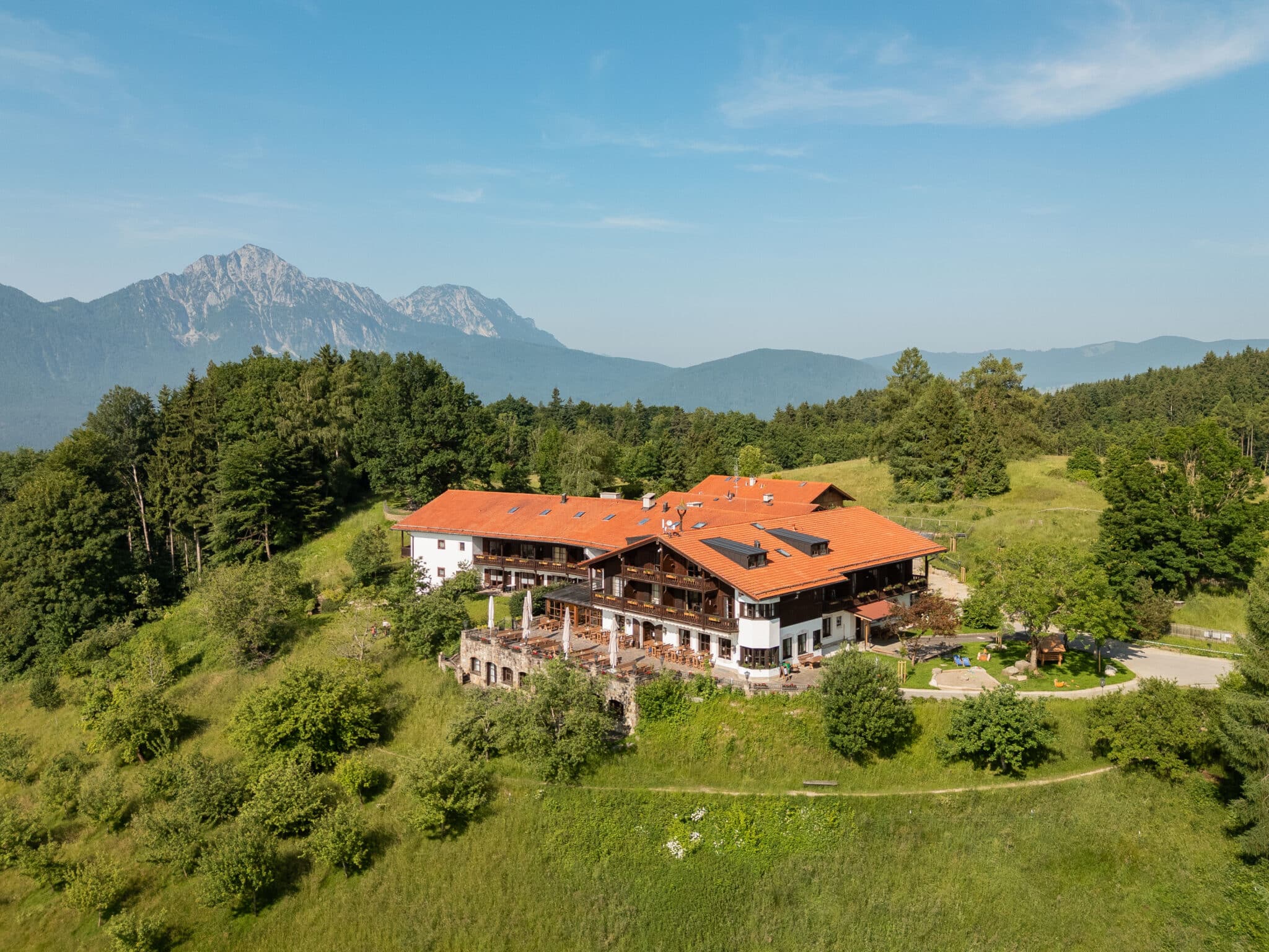 Berggasthof mit roten Ziegeldächern auf einer sonnigen, grasbewachsenen Anhöhe, umgeben von Bäumen und einer Außenterrasse, vor einem Panorama bewaldeter Hügel und markanter Berggipfel im Hintergrund.