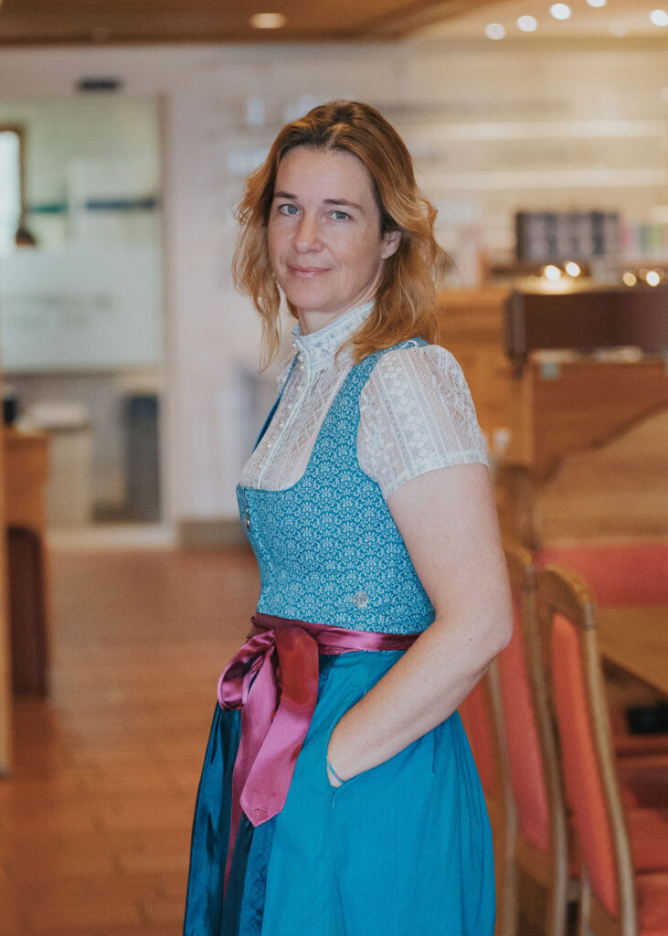 Frau mittleren Alters mit schulterlangem hellbraunem Haar in einem blauen Dirndl mit weißer Spitzenbluse und pinker Schürzenbinde, stehend in einem warm beleuchteten Gastraum mit Stühlen im Hintergrund und den Händen in den Taschen.