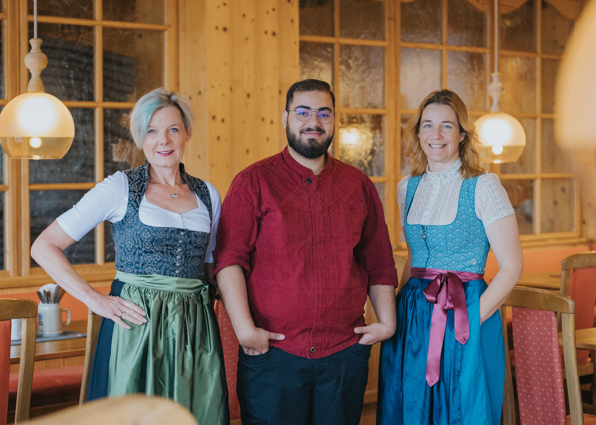 Drei lächelnde Personen stehen nebeneinander in einem warm beleuchteten, holzvertäfelten Gastraum; zwei Frauen tragen traditionelle Dirndl mit grüner bzw. blauer Schürze, ein Mann in rotem Hemd und Brille steht in der Mitte.