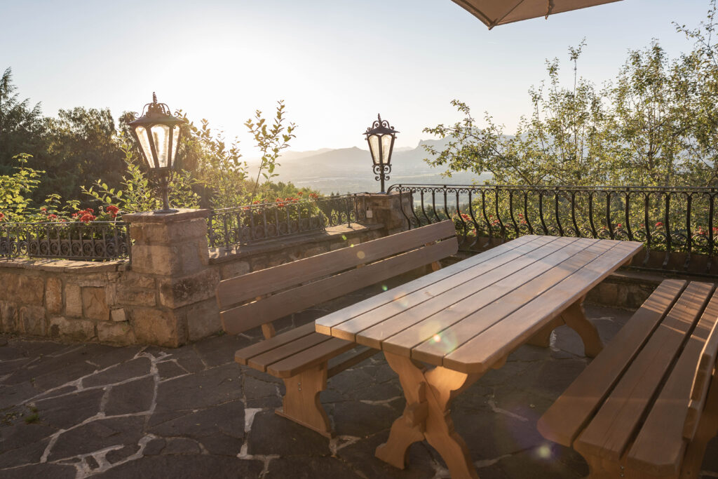 Terrasse bei Sonnenuntergang mit einer hölzernen Tisch‑und‑Bank‑Garnitur im Vordergrund, steinerner Brüstung mit schmiedeeisernem Geländer und dekorativen Laternen, Blumen und Bäumen sowie weitem Tal‑ und Bergpanorama im Hintergrund.
