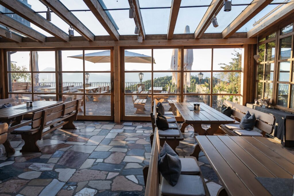 Gemütlicher, hell erleuchteter Wintergarten mit sichtbaren Holzbalken und Glasdach, mehreren langen Holztischen und -bänken mit Kissen auf einem mehrfarbigen Natursteinboden und großen Fenstern, die auf eine Terrasse mit Sonnenschirmen sowie eine entfernte See‑ und Berglandschaft blicken.
