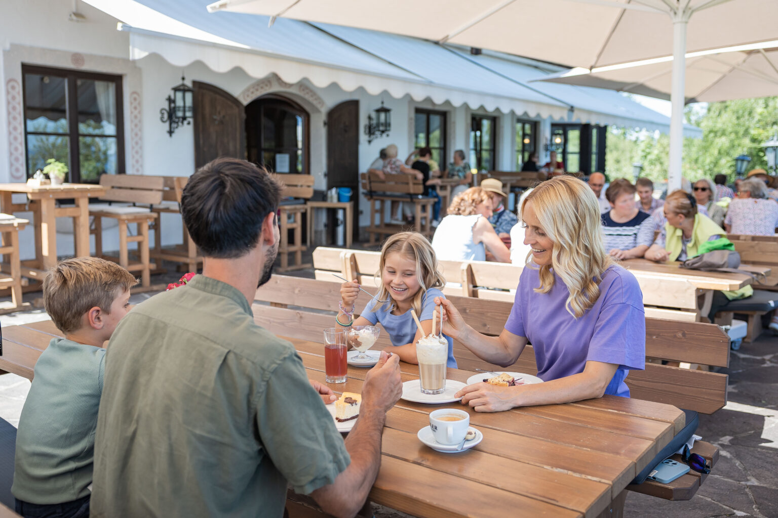 Familie (Mutter in lila Shirt, Vater mit dem Rücken zur Kamera und zwei Kinder) sitzt an einem hölzernen Außentisch eines Cafés unter Sonnenschirmen und genießt Eis, Kuchen und Kaffee vor einem weiß gestrichenen Haus mit Holzfenstern.