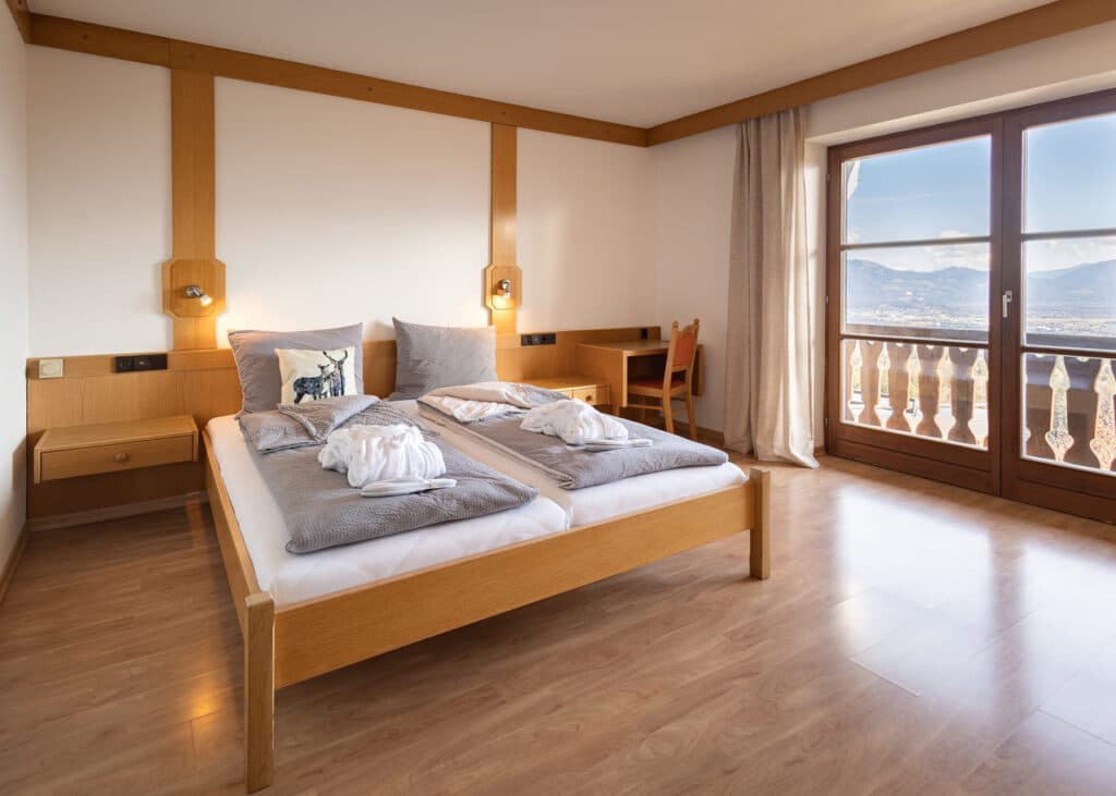 Helles, schlichtes Hotelzimmer mit zwei zusammengestellten Einzelbetten aus hellem Holz, grauen Bettdecken, dekorativem Kissen mit Hirschmotiv und aufgelegten Bademänteln, Holzkopfleiste mit Leselampen, kleinem Schreibtisch und großen Balkontüren, die auf Berge und Tal hinausblicken.
