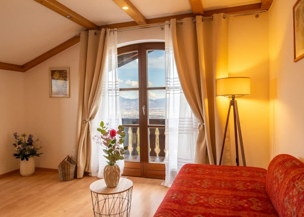 Gemütliches Wohnzimmer mit geschwungenem Holz-Balkontürfenster und Bergblick, hellen Vorhängen, einem kleinen Rundtisch mit Vase und Blumen im Vordergrund, rotem gemustertem Sofa rechts, Stehlampe und dekorativen Pflanzen in der Ecke.