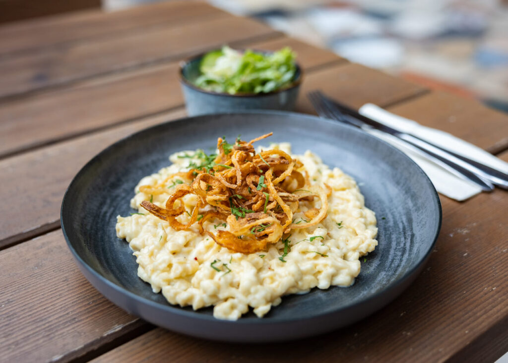 Schwarzer Teller mit cremigen Käsespätzle, obenauf knusprige Röstzwiebeln und gehackte Kräuter, neben Besteck auf einem Holztisch und einer kleinen Salatschüssel im unscharfen Hintergrund.