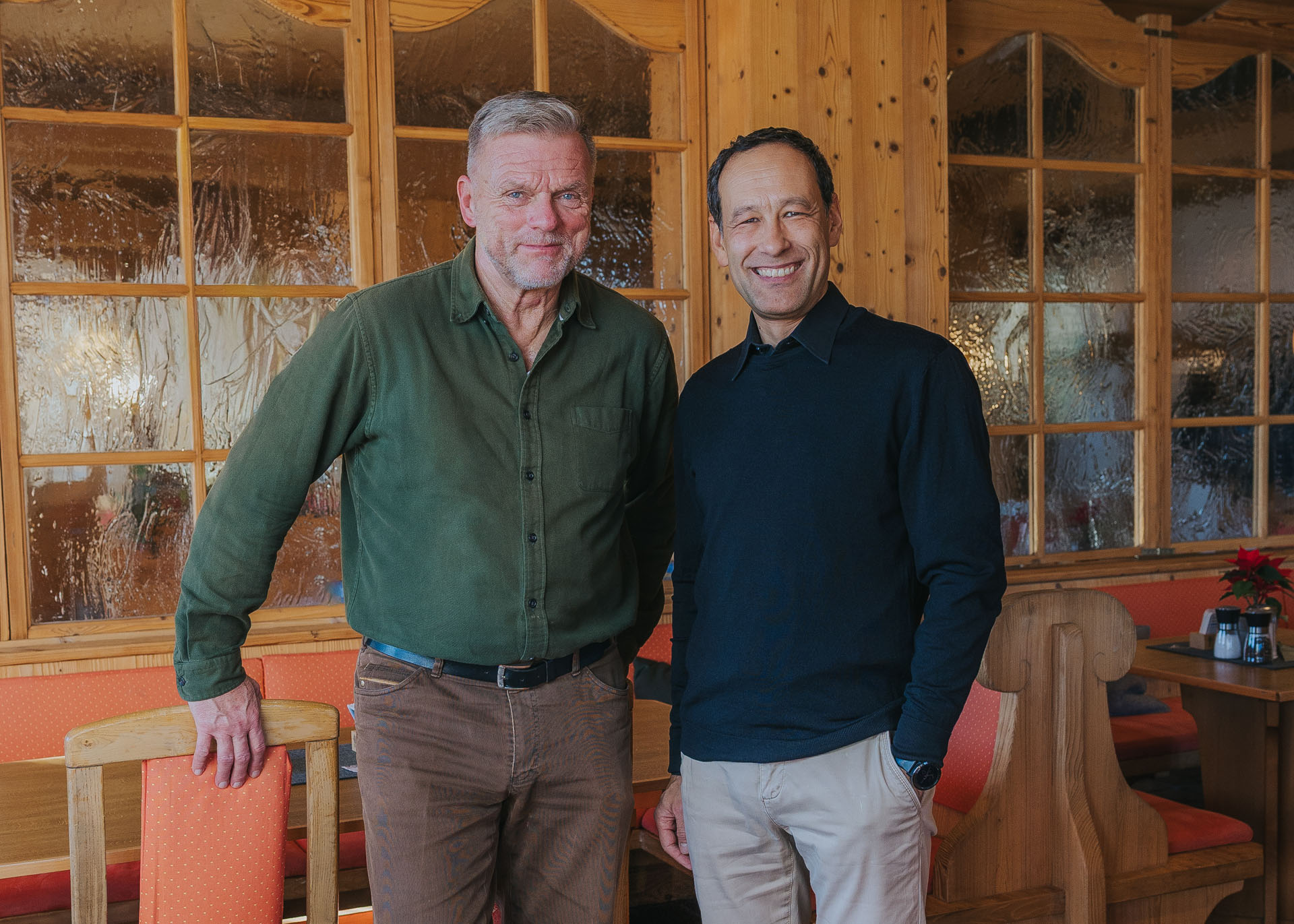 Zwei lächelnde Männer in legerer Kleidung stehen in einer holzvertäfelten Gaststube vor mattierten Fenstern.