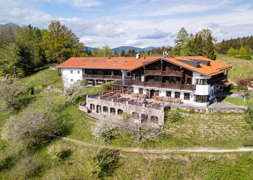 Alpenhotel mit rotem Ziegeldach, Holzbalkonen und großer Außenterrasse auf steiler, blühender Hangwiese vor Wald und Bergen.