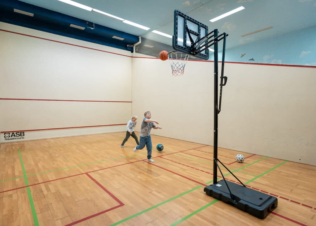 Zwei Jungen in einer Squash-Halle spielen Basketball: ein Junge wirft den Ball Richtung eines mobilen Korbs (Ball kurz vor dem Ring), der andere läuft hinterher; helles Parkett mit farbigen Spiellinien und ein Fußball am Rand.