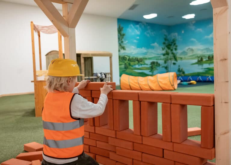 Kind baut mit großen roten Spielsteinen eine Mauer in einer Indoor-Spielhalle